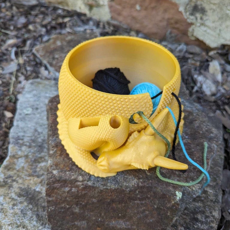 May include: Yellow ceramic yarn bowl shaped like a dragon with a blue, green, and black yarn ball inside.