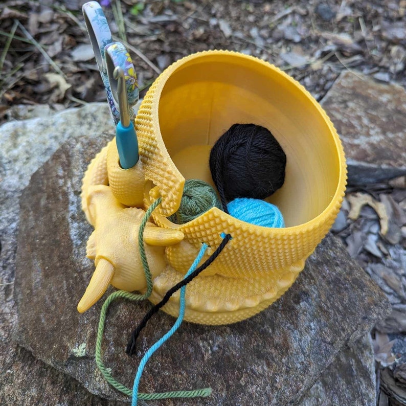 May include: A yellow ceramic yarn bowl shaped like a turtle with a blue crochet hook and three balls of yarn inside: black, teal, and green.