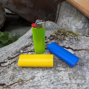May include: A green lighter with a red top and silver metal accents, next to a yellow and a blue lighter sleeve. The items are on a weathered, gray wooden surface.
