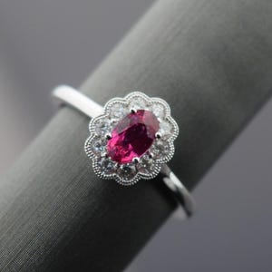 May include: A silver ring with a central oval ruby, encircled by a halo of smaller, clear gemstones. The ring has a delicate, scalloped edge and a thin, polished band. The ruby is a deep, rich red.
