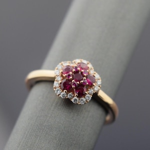 May include: A gold ring with a floral cluster of red gemstones, encircled by a halo of smaller, clear stones. The ring sits on a gold band, presenting a classic and elegant design. The ring's design is timeless.