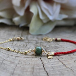 Op de afbeelding: Een goudkleurige armband met een groene edelsteen en een rood koord. De armband heeft een delicate ketting met kleine gouden kralen en een centrale groene steen. Het rode koord zit aan de andere kant.