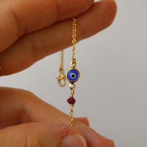 May include: A gold-toned chain bracelet featuring a small, circular blue and white evil eye charm and a single, small, faceted dark pink gemstone bead. The delicate metal links connect the charms in a vertical arrangement on the thin chain.
