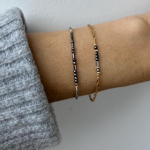 May include: Two delicate bracelets. One is silver with black beads in a vertical line. The other is gold with small black beads. The bracelets are worn on a wrist, with a gray sweater sleeve in the background.