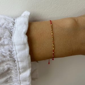Puede incluir: Una delicada pulsera de hilo rojo con pequeñas cuentas doradas. La pulsera se lleva en una muñeca sobre un fondo blanco. Las cuentas doradas están espaciadas uniformemente a lo largo del hilo rojo, creando un diseño minimalista y elegante.