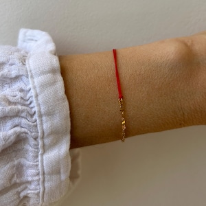 Puede incluir: Una pulsera de hilo rojo con una delicada extensión de cadena dorada. La pulsera se lleva en la muñeca, con una manga de camisa blanca visible en el fondo. Una joya sencilla y elegante.