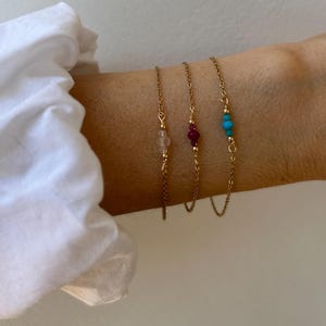 Könnte beinhalten: Drei zarte goldene Kettenarmbänder, jedes mit einem einzelnen Edelstein. Eines hat einen rosa Stein, ein anderes ein tiefes Rot und das dritte Türkis. Die Armbänder werden an einem Handgelenk vor neutralem Hintergrund gezeigt.