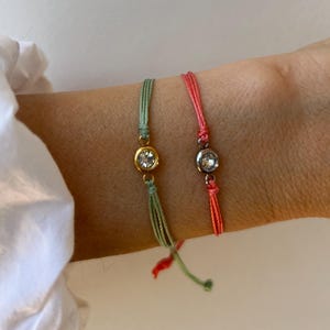 May include: Two string bracelets featuring round, clear faceted stones. One bracelet has a sage green cord with a gold-toned setting, while the other has a coral pink cord with a silver-toned setting. Both bracelets are adjustable with loose ends.