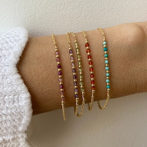 Può includere: Cinque braccialetti a catena d'oro con perline di pietre preziose di diversi colori. I braccialetti sono disposti in ordine arcobaleno, dal viola al turchese.