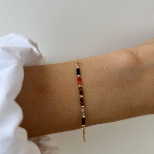 Könnte beinhalten: Ein zartes Goldkettenarmband mit einer Reihe kleiner, mehrfarbiger Edelsteinperlen in den Farben Schwarz, Rot, Braun und durchscheinendem Grau, die mit winzigen goldfarbenen Metallzwischenstücken entlang der dünnen Kette angeordnet sind.