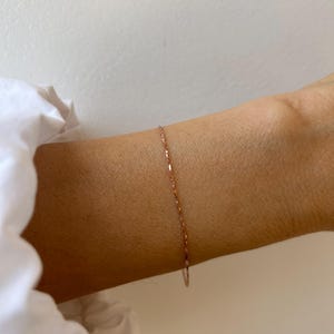 Puede incluir: Una delicada pulsera de cadena en oro rosa con pequeños eslabones planos y reflectantes que crean un sutil efecto brillante. La fina cadena de metal descansa sobre la piel, mostrando un acabado pulido y un diseño sencillo y continuo.