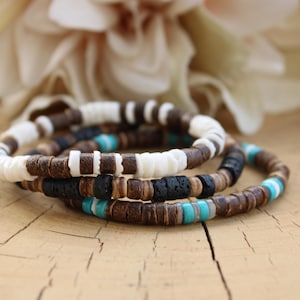 Könnte beinhalten: Ein Stapel aus drei Armbändern, die aus kleinen, flachen, runden Perlen bestehen. Die Perlen sind in den Farben Braun, Weiß, Schwarz und Türkis gehalten. Die Armbänder zeigen eine Mischung aus glatten, strukturierten und porösen Oberflächen.