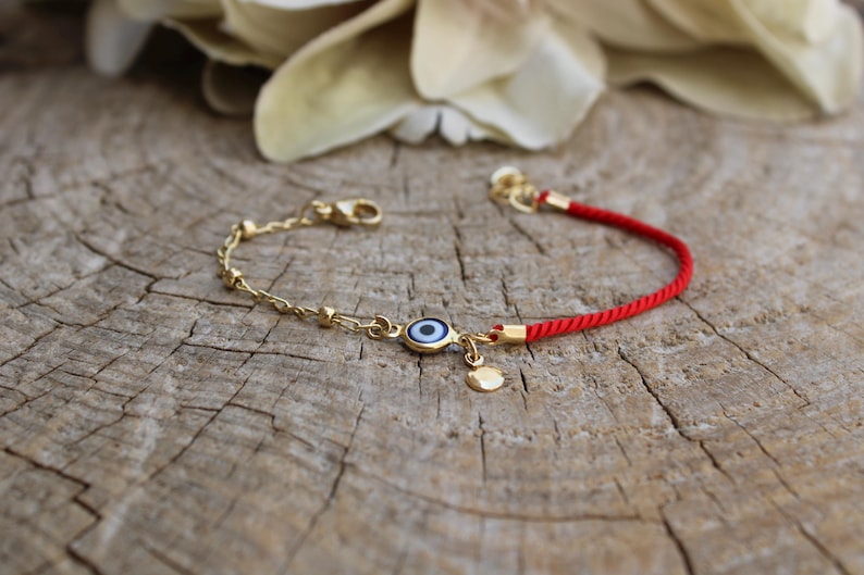 May include: A red string bracelet with a gold chain and a blue evil eye charm. The charm has a white centre and a gold rim.
