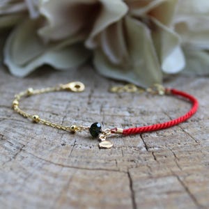 May include: A red string bracelet with a gold chain and a black bead. The bracelet has a small gold charm.