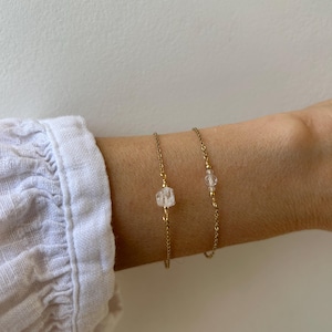May include: Two delicate gold-toned chain bracelets, each featuring a single small, clear, irregularly shaped stone bead centered on the chain. The bracelets are thin and minimalist in construction, showcasing a simple link design with metallic gold finish.