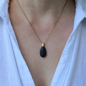 Könnte beinhalten: Goldfarbene Halskette mit einem schwarzen, tropfenförmigen Anhänger. Der Anhänger hat eine strukturierte Oberfläche und hängt an einer feinen Kette. Die Halskette wird über einem weißen Hemd getragen.