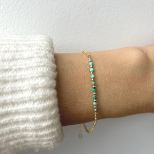 Könnte beinhalten: Ein zartes Armband aus goldener Kette, verziert mit kleinen, runden türkisfarbenen Perlen. Das Armband wird an einem Handgelenk gezeigt, mit einem weißen Ärmel im Hintergrund. Das Design des Armbands ist schlicht und elegant.