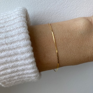 Puede incluir: Una delicada pulsera de cadena dorada con un diseño de eslabones planos y reflectantes que descansa sobre la piel. La pulsera es fina y pulida, captando la luz con su superficie metálica, creando una línea dorada sutil y continua alrededor de la muñeca.