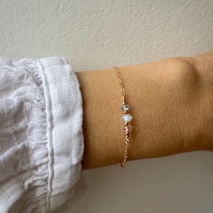 May include: A delicate gold chain bracelet with three small beads: a light pink, a clear iridescent, and a light green. The bracelet is on a person's wrist.
