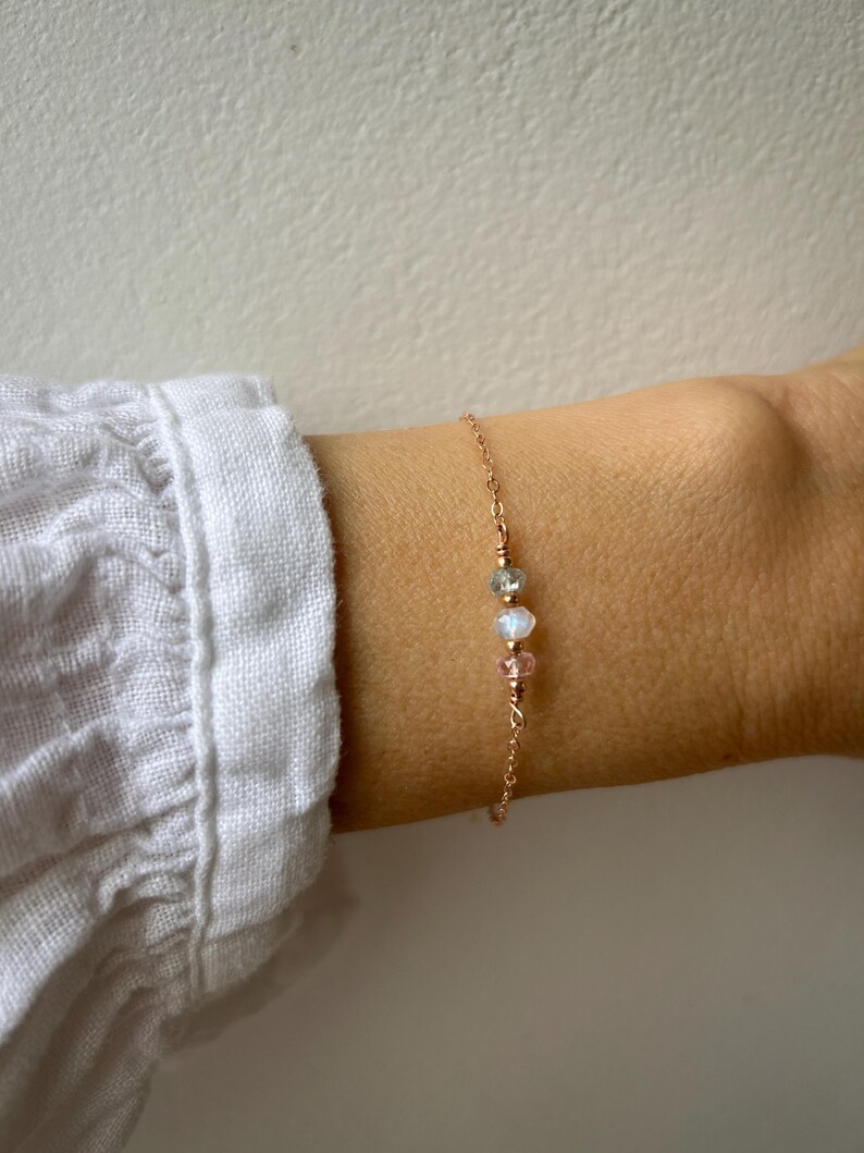 May include: A delicate gold chain bracelet with three small beads: a light pink, a clear iridescent, and a light green. The bracelet is on a person's wrist.
