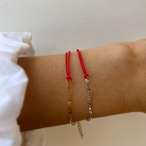 Può includere: Due braccialetti delicati con segmenti di cordino rosso sottile collegati a catene metalliche. Un braccialetto ha una catena color oro, mentre l'altro ha una catena color argento. Entrambi includono una piccola catenella di estensione regolabile.