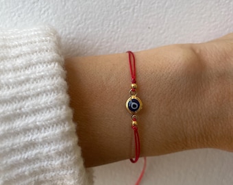 May include: A red string bracelet featuring a central circular charm with a blue, white, and black evil eye design. The charm is encased in a gold-toned metal frame and is flanked by two small gold-toned beads on a thin, adjustable red cord.