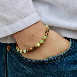 Puede incluir: Un brazalete hecho de cuentas de madera de color verde claro y marrón. El brazalete se lleva en la muñeca.