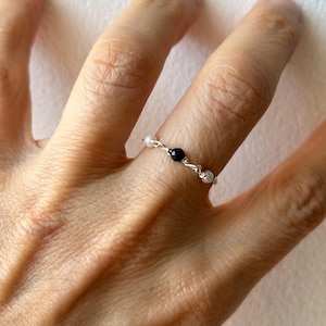 Op de afbeelding: Een delicate ring met een dunne, zilverkleurige draadband, geaccentueerd met een enkele zwarte kraal in het midden, geflankeerd door twee kleine witte kralen. De draad is tussen de kralen gedraaid, wat een eenvoudig, lineair ontwerp vormt.