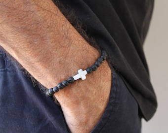 May include: A stretch bracelet featuring small, round, black porous lava stone beads arranged in a continuous loop, accented by a single, smooth, white cross-shaped bead positioned centrally along the strand of dark, textured volcanic rock beads.