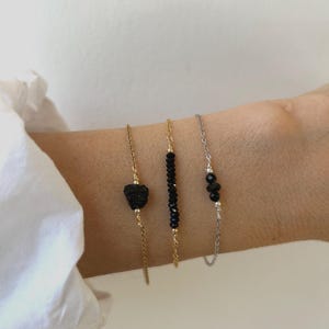 Könnte beinhalten: Drei zarte Armbänder, die zusammen getragen werden. Das erste ist eine goldene Kette mit einem einzelnen rohen schwarzen Stein. Das zweite ist eine goldene Kette mit einer Reihe kleiner schwarzer Facettenperlen. Das dritte ist eine silberne Kette mit zwei kleinen schwarzen Perlen.