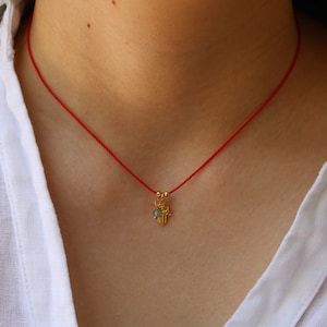 Puede incluir: Un collar de cuerda roja con un dije de mano de hamsa de oro y una piedra preciosa azul. El collar lo lleva una persona con una camisa blanca.