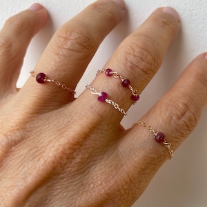 May include: Three delicate gold rings with small ruby red stones. The rings are worn on a finger.