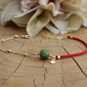 May include: A bracelet featuring a gold-tone chain on one side and a red cord on the other. A single round green stone bead is centered, accented by a small gold-tone charm and a tiny red bead. The bracelet includes a gold-tone lobster clasp.