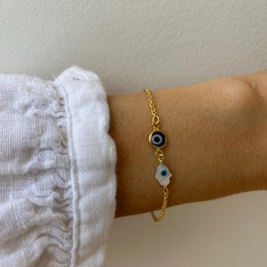 Può includere: Un bracciale a catena color oro con due ciondoli: una perla circolare blu e bianca a forma di occhio di Allah e un ciondolo a forma di mano di Fatima in madreperla bianca con un piccolo occhio blu al centro. La sottile catena unisce i ciondoli.