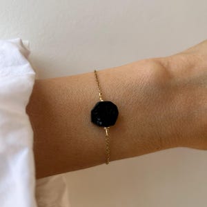 Op de afbeelding: Een verfijnde goudkleurige kettingarmband met een enkele, gefacetteerde, achthoekige zwarte steen in het midden. De steen is aan beide zijden vastgezet met dunne goudkleurige draadwikkelingen die verbonden zijn met de fijne schakelketting.