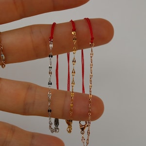 Può includere: Primo piano di diverse collane delicate. Una è una catena d'oro con piccole perle, mentre le altre sono sottili catene in argento, oro e oro rosa. Ogni catena è attaccata a un cordino rosso.