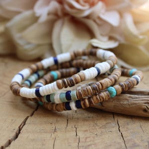 Peut inclure: Un empilement de trois bracelets composés de petites perles rondes en bois brun alternées avec des perles en forme de disque de couleur blanche, bleu foncé et turquoise. Les bracelets sont disposés en cercle, révélant des textures naturelles.