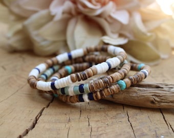 Pode incluir: Uma pilha de três pulseiras feitas de pequenas contas redondas de cor castanha semelhante a madeira, intercaladas com contas em forma de disco brancas, azul-escuras e turquesa. As pulseiras estão dispostas em círculo, exibindo texturas.