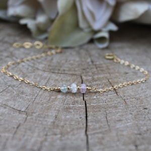 May include: A delicate gold chain bracelet with three small gemstone beads. The beads are light blue, white, and pink.