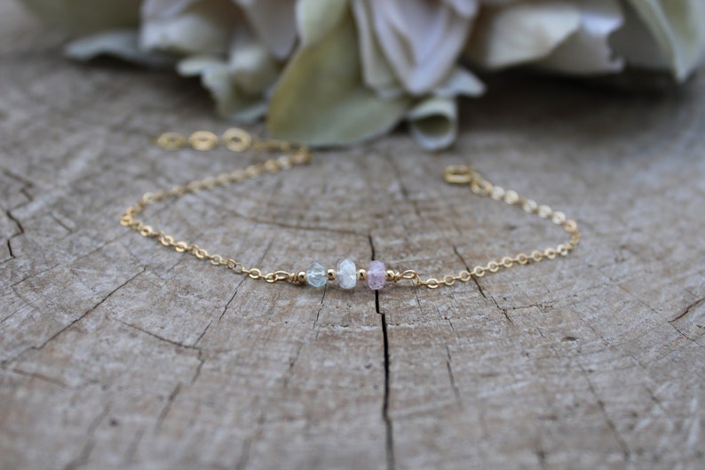 May include: A delicate gold chain bracelet with three small gemstone beads. The beads are light blue, white, and pink.