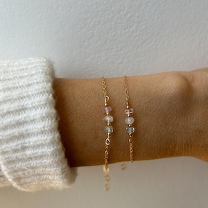 May include: Two gold chain bracelets with small pink and blue gemstone beads. The bracelets are layered on a person's wrist.