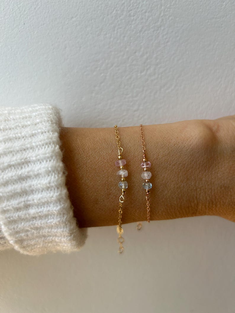 May include: Two gold chain bracelets with small pink and blue gemstone beads. The bracelets are layered on a person's wrist.