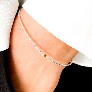 Puede incluir: Un delicado collar de cadena de plata con un pequeño acento de cuentas doradas. El collar lo lleva una persona con una camisa blanca y una blusa negra.