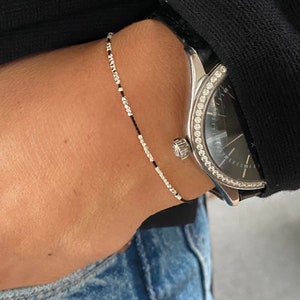 Può includere: Un braccialetto di perline d'argento con un cordoncino nero sul polso di una persona. Il braccialetto è indossato con un orologio con una lunetta di diamanti.