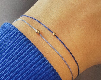 May include: Two bracelets on a wrist. The bracelets are made of thin blue and purple cords with small gold beads at the ends.