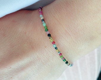 May include: A colourful gemstone bracelet with small beads in shades of pink, green, blue, and gold. The beads are strung on a thin cord. The bracelet is worn on a wrist, highlighting its vibrant colours and delicate design. A simple and elegant piece of jewellery.
