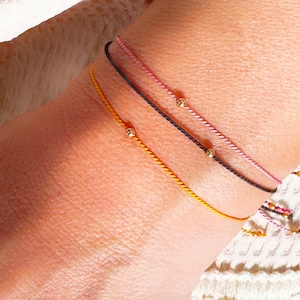 Puede incluir: Tres pulseras con cuentas doradas en la muñeca de una persona. Las pulseras son rosa, negra y amarilla.