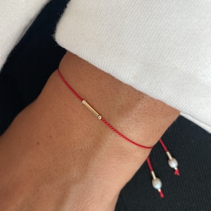 Peut inclure: Un bracelet en cordon rouge avec un charme en forme de barre dorée et deux petits accents de perles blanches.