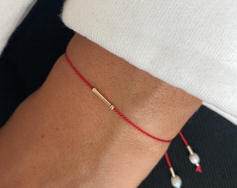 May include: A red string bracelet with a gold bar charm and two small white pearl accents.