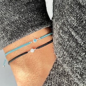 May include: Three delicate bracelets. One is black, one is light blue, and the other is a darker blue. Each bracelet has a small, round, silver-colored charm with a single gemstone. The bracelets are worn on a wrist.